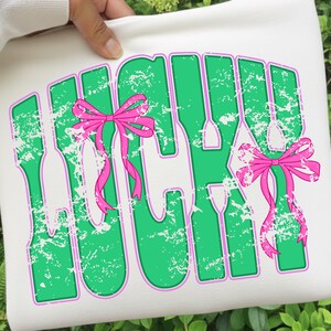May include: White tote bag with the word "LUCKY" in green, outlined in purple, with a distressed texture. Pink bows are attached to the letters. The bag is held by a person.