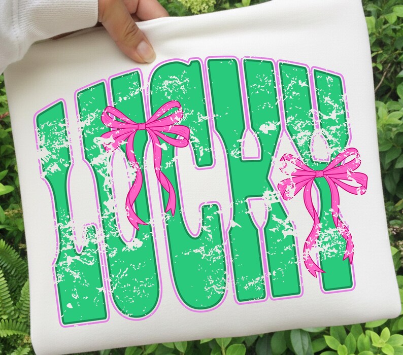 May include: White tote bag with the word "LUCKY" in green, outlined in purple, with a distressed texture. Pink bows are attached to the letters. The bag is held by a person.