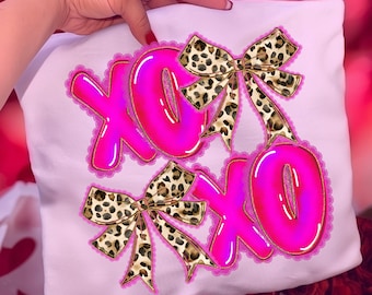 May include: A white pillow with a bright pink "XOXO" design. The letters are outlined in gold and have a bubble letter style. Two leopard print bows are placed above and below the letters. The design is surrounded by a pink lace trim.