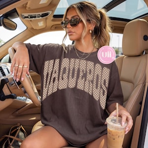 May include: A dark brown short-sleeve t-shirt featuring the word VAQUEROS printed in large, arched, beige letters with a white polka dot pattern. The shirt has a crew neckline and a loose, relaxed fit, worn while sitting in a vehicle interior.