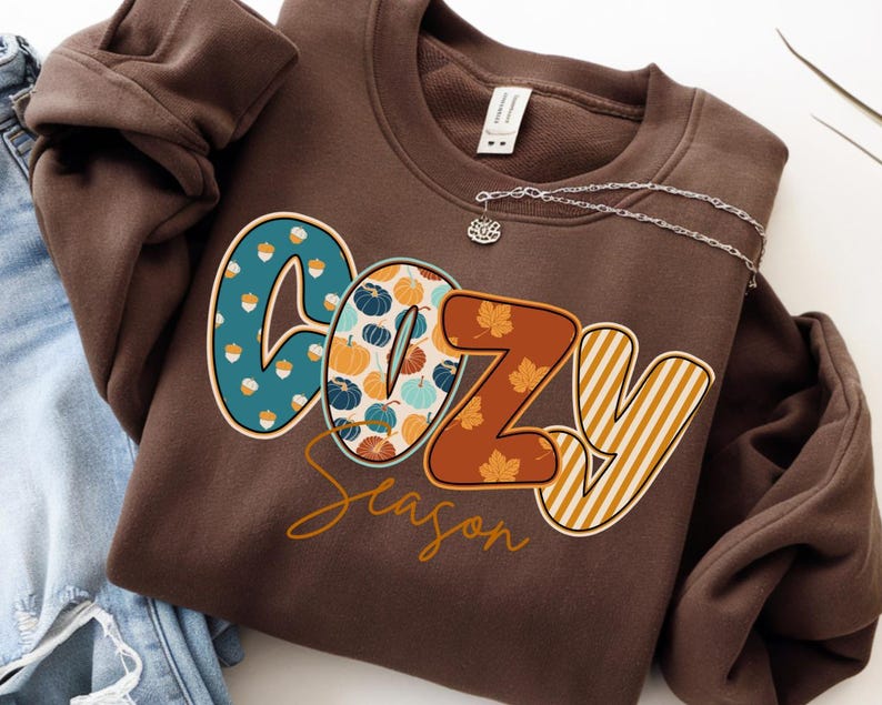 May include: A brown sweatshirt with the words "COZY Season" in a fall-themed design. The letter "C" is teal with acorn graphics, "O" has pumpkin graphics, "Z" has leaf graphics, and "Y" has vertical stripes. A silver necklace is attached.