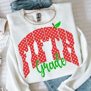 May include: A white crewneck sweatshirt featuring a red graphic with white polka dots shaped like an apple. The graphic contains the text "FIFTH" in large block letters and the word "Grade" in green cursive script below the apple design.