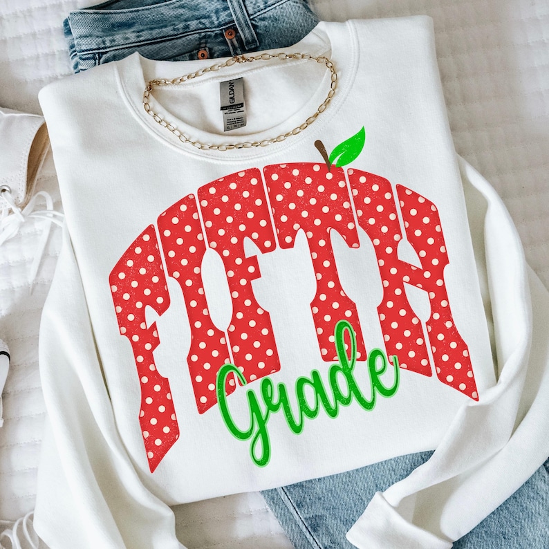 May include: A white crewneck sweatshirt featuring a red graphic with white polka dots shaped like an apple. The graphic contains the text "FIFTH" in large block letters and the word "Grade" in green cursive script below the apple design.