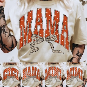 May include: A cream-colored t-shirt featuring the word MAMA in large, orange, white-polka-dotted letters. A leopard-print bow graphic is layered over the bottom of the text. The design is repeated in smaller variations with the words GIGI, NANA, MINI, and MIMI.