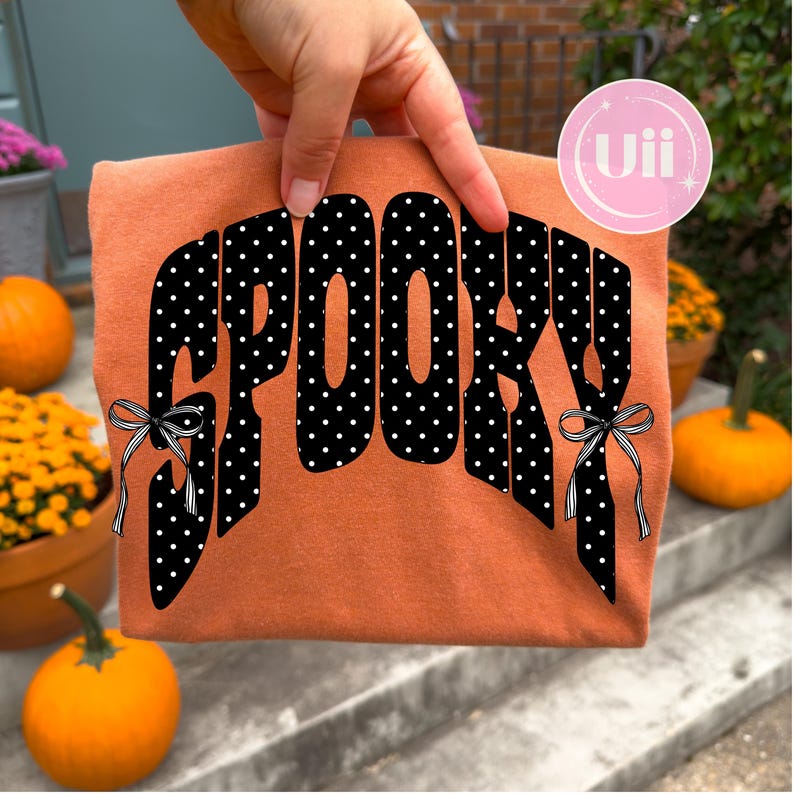 K&ouml;nnte beinhalten: Ein orangefarbenes T-Shirt mit dem Wort SPOOKY in gro&szlig;en schwarzen Buchstaben mit wei&szlig;en Punkten. Die Buchstaben S und Y sind mit kleinen schwarz-wei&szlig; gestreiften Schleifen verziert. Der Schriftzug ist mittig auf dem Stoff gebogen.
