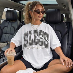 May include: White t-shirt with the text "BLESS your heart" in a black polka dot design. The shirt is worn with black shorts and white sneakers. The person is wearing sunglasses and jewelry.