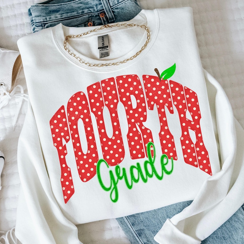 May include: A white long-sleeve sweatshirt featuring a large graphic that reads "FOURTH Grade" in red with white polka dots. The word "FOURTH" is in a bold, blocky font, and "Grade" is in a green cursive script with a small green leaf icon.