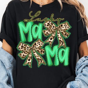 May include: Black t-shirt with a green and gold design. The design features the words "Lucky Mama" in gold script above two green "Ma" letters. Two leopard print bows are also included in the design.