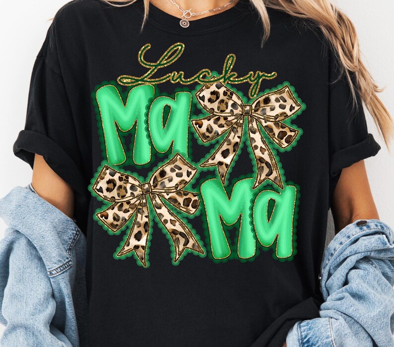 May include: Black t-shirt with a green and gold design. The design features the words "Lucky Mama" in gold script above two green "Ma" letters. Two leopard print bows are also included in the design.