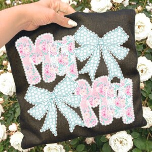 May include: Black pillow with the word "MOM" in pink floral letters and light blue polka dot bows. The letters have a white scalloped edge. The pillow is held by a hand.