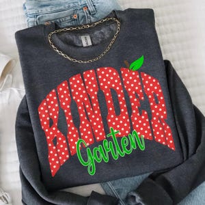 May include: A dark charcoal gray crewneck sweatshirt featuring the text "Kinder Garten" in a bold, red, white-polka-dotted font. A small green leaf graphic sits atop the letter "i". A thin gold-toned chain necklace rests on the collar.
