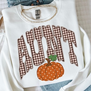 May include: A white crewneck sweatshirt featuring the word AUNT in large, brown and white gingham plaid letters. Below the text is an orange pumpkin with white polka dots and a green stem. A gold-toned chain necklace rests on the collar.