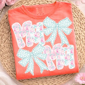 May include: Coral-colored t-shirt with the word "MOM" in a floral pattern, outlined with white lace. Two light blue polka-dot bows are above and below the word. The shirt is folded on a woven mat.