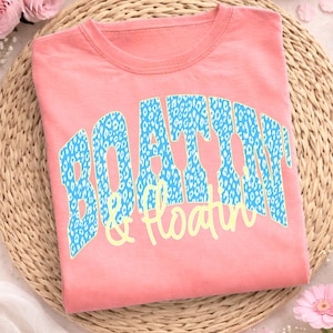 May include: A coral t-shirt featuring the words "BOATTIN" and "& Floatin" in a blue leopard print with a yellow outline. The shirt is folded on a woven mat. A casual summer top.