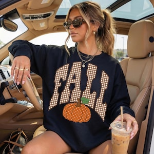 Peut inclure: Un sweat-shirt noir &agrave; manches longues avec le mot FALL &eacute;crit en motif vichy marron et blanc sur la poitrine. Sous le texte se trouve un graphique repr&eacute;sentant une citrouille orange &agrave; pois blancs avec une tige verte. Col rond classique.
