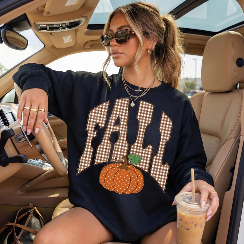 Peut inclure: Un sweat-shirt noir &agrave; manches longues avec le mot FALL &eacute;crit en motif vichy marron et blanc sur la poitrine. Sous le texte se trouve un graphique repr&eacute;sentant une citrouille orange &agrave; pois blancs avec une tige verte. Col rond classique.