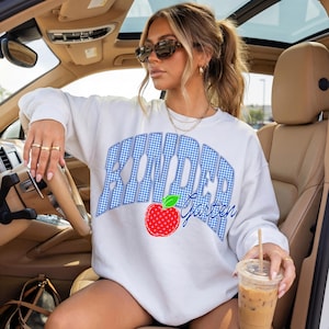 May include: White sweatshirt with the text "Kindergarten" in a blue gingham design. A red apple with a green leaf is below the text. The sweatshirt is a casual style.