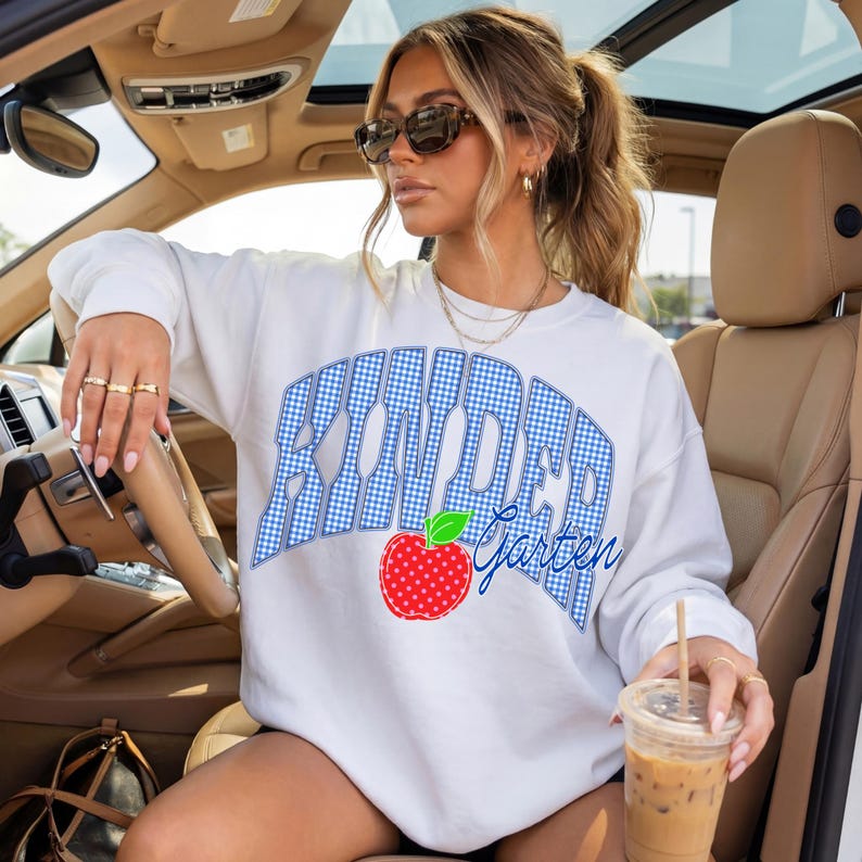 May include: White sweatshirt with the text "Kindergarten" in a blue gingham design. A red apple with a green leaf is below the text. The sweatshirt is a casual style.