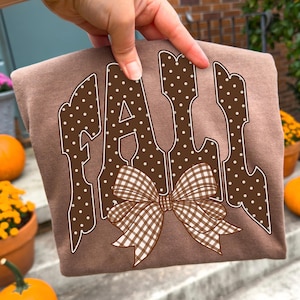 May include: A brown t-shirt featuring the word FALL in large, blocky, dark brown letters with white polka dots. A brown and white gingham patterned bow is positioned over the bottom of the letters. The design is centered on the front of the shirt.