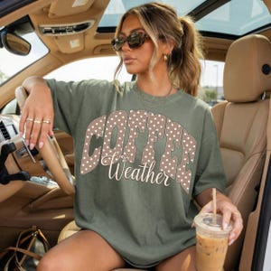 May include: Olive green t-shirt with the words "COFFEE Weather" in a polka-dot design. The shirt is worn by a person in a car, holding a coffee drink.