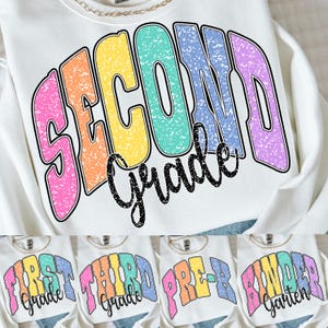 May include: A white crewneck sweatshirt featuring a multi-coloured graphic with a distressed texture. The text reads SECOND Grade in large, arched, pastel-coloured letters with a black outline, layered over a smaller black cursive script.