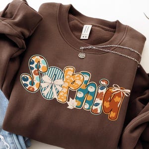 May include: A brown sweatshirt with the name "Sophie" in a colorful, fall-themed design. The letters feature pumpkins, leaves, and stripes in shades of orange, teal, and white. A silver necklace with a small charm is pinned to the sweatshirt.