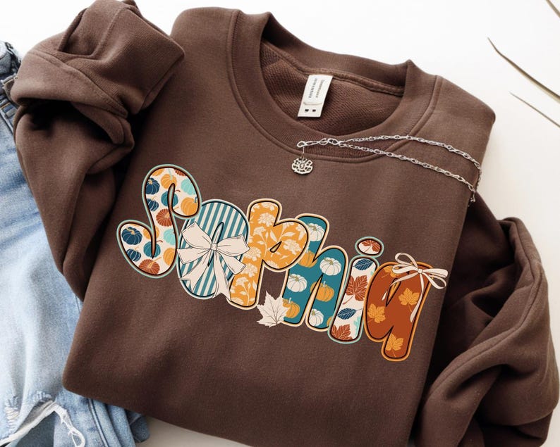 May include: A brown sweatshirt with the name "Sophie" in a colorful, fall-themed design. The letters feature pumpkins, leaves, and stripes in shades of orange, teal, and white. A silver necklace with a small charm is pinned to the sweatshirt.