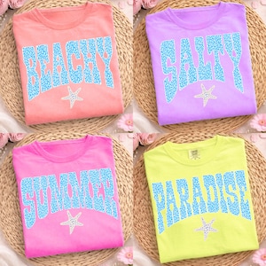 May include: Four brightly coloured t-shirts: peach, lilac, pink, and lime. Each shirt features a different word in blue, textured lettering: "BEACHY", "SALTY", "SUMMER", and "PARADISE". A white starfish is below each word. The shirts are on woven mats.