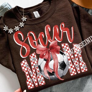 May include: A brown sweatshirt with the words "Soccer Mom" in red script and checkered letters. A soccer ball with a red bow is in the center of the design. A floral necklace is draped on the sweatshirt.