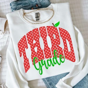 May include: A white crewneck sweatshirt featuring a graphic with the text "Third Grade" in red with white polka dots. The word "Third" is in a bold, arched font, and "Grade" is in a bright green script font with a small green apple leaf.