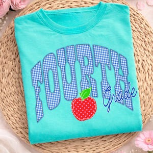 May include: A turquoise t-shirt with the words "FOURTH Grade" in a blue gingham pattern. A red apple with white polka dots and a green leaf is below the text. The shirt is folded on a woven mat.