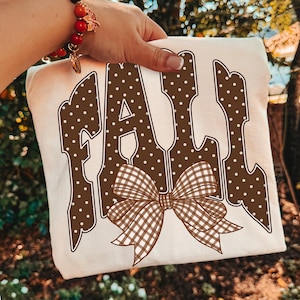 May include: A cream-colored t-shirt featuring the word FALL in large, brown, white-polka-dotted letters. A brown and white gingham patterned bow is positioned over the bottom of the letters, centered on the front of the garment.