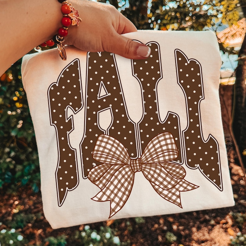 May include: A cream-colored t-shirt featuring the word FALL in large, brown, white-polka-dotted letters. A brown and white gingham patterned bow is positioned over the bottom of the letters, centered on the front of the garment.