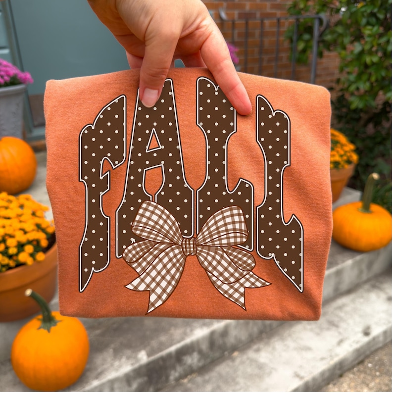 May include: An orange t-shirt featuring the word FALL in large, brown, white-polka-dotted block letters. A brown and white gingham patterned bow is centered over the bottom of the letters. The design is printed on the front of the fabric.