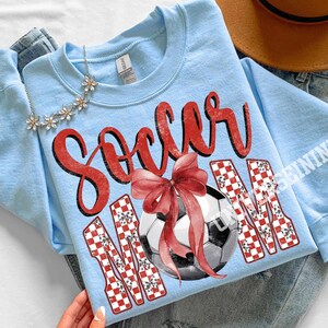May include: Light blue sweatshirt with the words "Soccer Mom" in red script and a soccer ball with a red bow. The letters "MOM" are in a red and white checkered pattern. A silver necklace with flower accents is at the top.