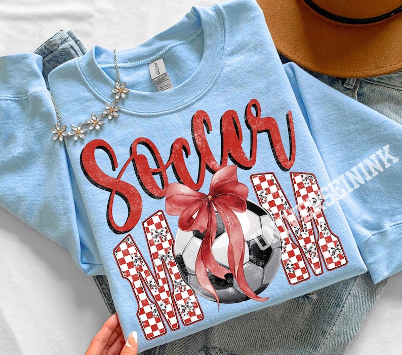 May include: Light blue sweatshirt with the words "Soccer Mom" in red script and a soccer ball with a red bow. The letters "MOM" are in a red and white checkered pattern. A silver necklace with flower accents is at the top.