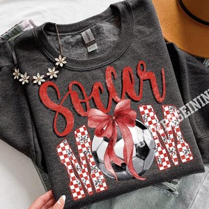 May include: Dark gray sweatshirt with the words "Soccer Mom" in red script and a soccer ball graphic. The word "MOM" is in a red and white checkered pattern. A red bow is over the soccer ball.