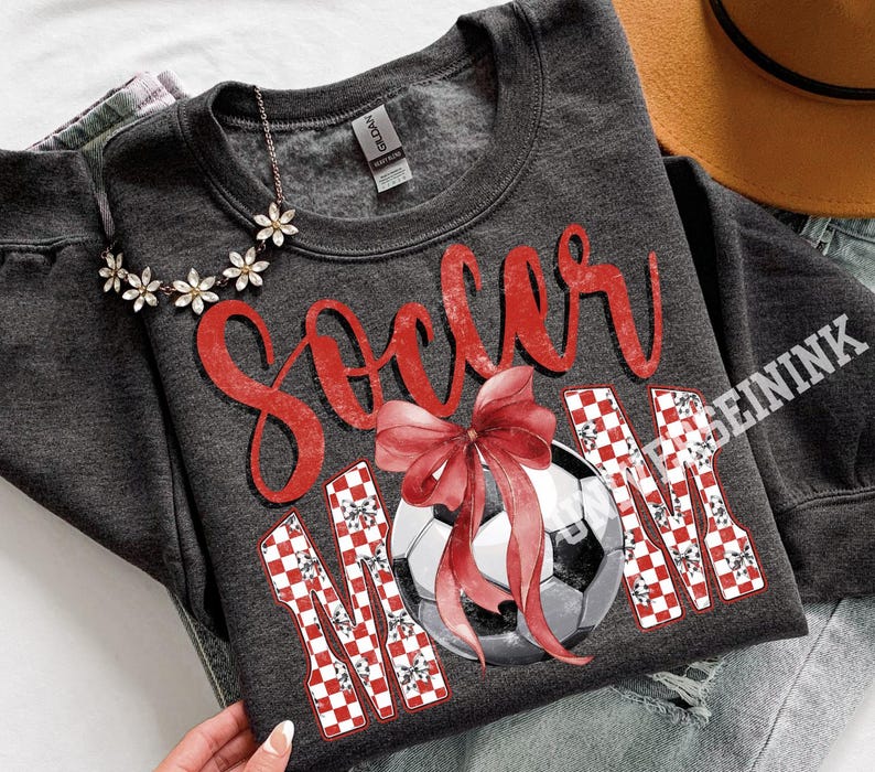 May include: Dark gray sweatshirt with the words "Soccer Mom" in red script and a soccer ball graphic. The word "MOM" is in a red and white checkered pattern. A red bow is over the soccer ball.