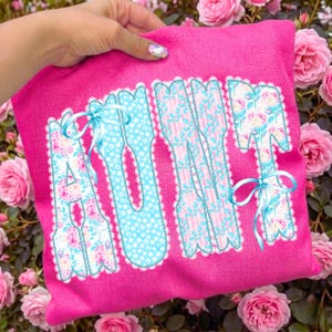 Puede incluir: Un cojín rosa con la palabra "AUNT" aplicada en un estampado floral y de lunares. Las letras están delineadas con encaje blanco y atadas con cintas azules. El cojín se sostiene frente a un fondo de rosas rosas.