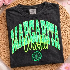 May include: Dark gray t-shirt with the words "MARGARITA Weather" in neon green. The word "MARGARITA" has a leopard print design. A lime graphic is below the text. The shirt is on a woven surface.