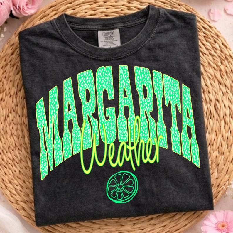May include: Dark gray t-shirt with the words "MARGARITA Weather" in neon green. The word "MARGARITA" has a leopard print design. A lime graphic is below the text. The shirt is on a woven surface.