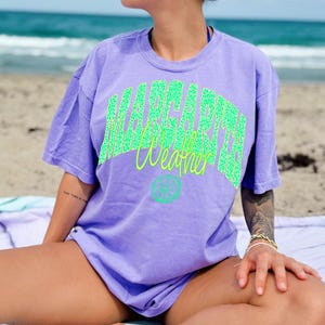 May include: A lilac-colored t-shirt with the words "Margarita Weather" in a green leopard print design and a lime graphic. The shirt is short-sleeved and has a relaxed fit. The image is taken on a beach.