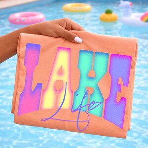May include: Peach-colored t-shirt with the words "LAKE life" in a colorful, gradient font. The shirt is held up against a backdrop of a swimming pool with inflatable toys.