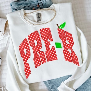 May include: A white long-sleeve sweatshirt featuring the text PRE-K in large, red, white-polka-dotted letters. The letter K is stylized with a green leaf at the top and a small green square accent. A gold-toned chain necklace rests at the neckline.
