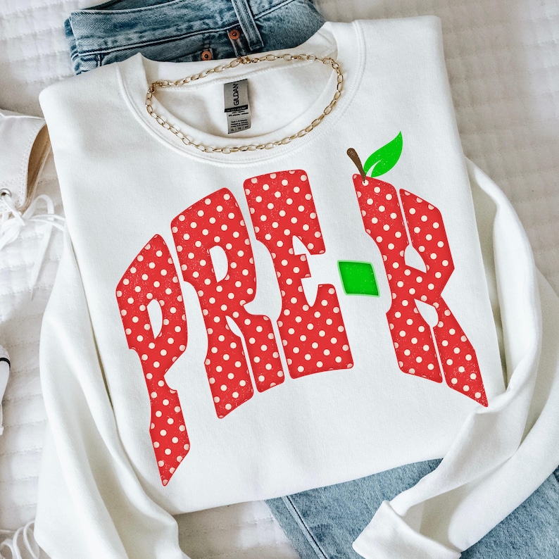 May include: A white long-sleeve sweatshirt featuring the text PRE-K in large, red, white-polka-dotted letters. The letter K is stylized with a green leaf at the top and a small green square accent. A gold-toned chain necklace rests at the neckline.