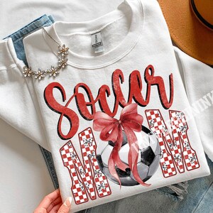May include: White sweatshirt with the words "Soccer Mom" in red script and checkered letters. A watercolor soccer ball with a red bow is in the center. The sweatshirt is folded on top of blue jeans.