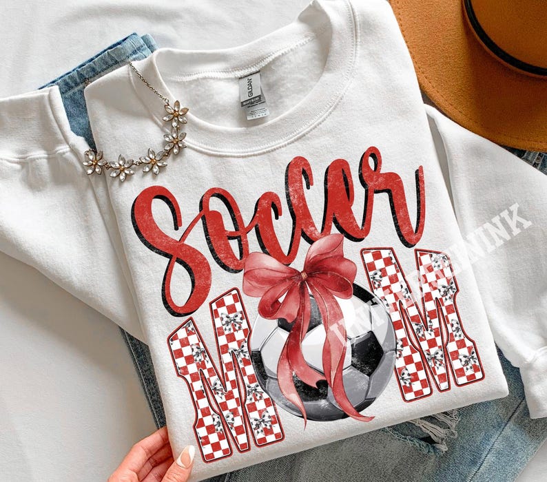 May include: White sweatshirt with the words "Soccer Mom" in red script and checkered letters. A watercolor soccer ball with a red bow is in the center. The sweatshirt is folded on top of blue jeans.