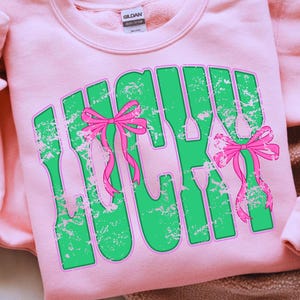 May include: A light pink sweatshirt with the word "LUCKY" in green, outlined in pink, and distressed. Pink bows are incorporated into the design. The sweatshirt is made of a soft material.