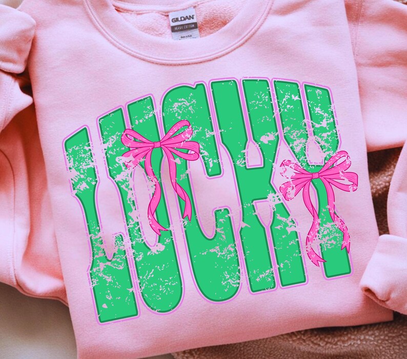 May include: A light pink sweatshirt with the word "LUCKY" in green, outlined in pink, and distressed. Pink bows are incorporated into the design. The sweatshirt is made of a soft material.