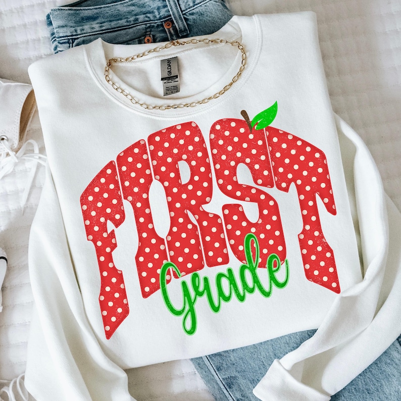May include: A white crewneck sweatshirt featuring a large red graphic with white polka dots that spells out the word FIRST in a bold, blocky font. Below this, the word Grade is written in a bright green cursive script with a small green leaf.